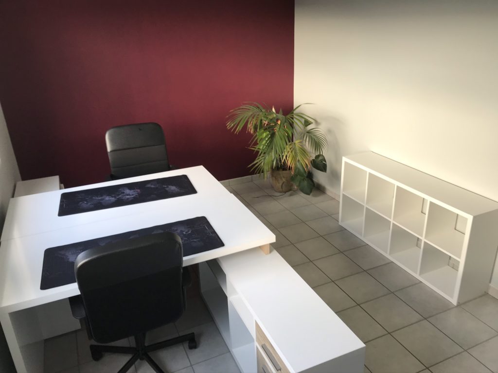 2 bureaux blancs. 2 chaises noires. une plante. un petit meuble blanc. un mur rouge. un mur blanc 