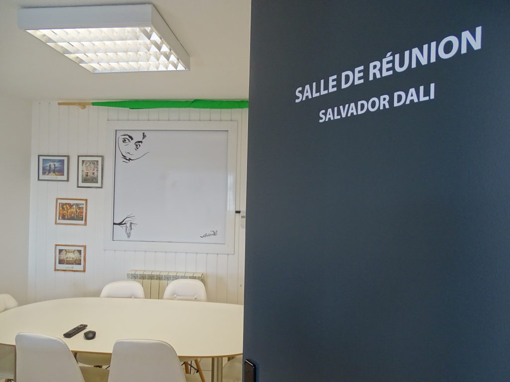 salle de réunion Saalvador dali avec table blanche, chaises blanches, tableau blanc, fond vert plié enroulé en haut du tableau.